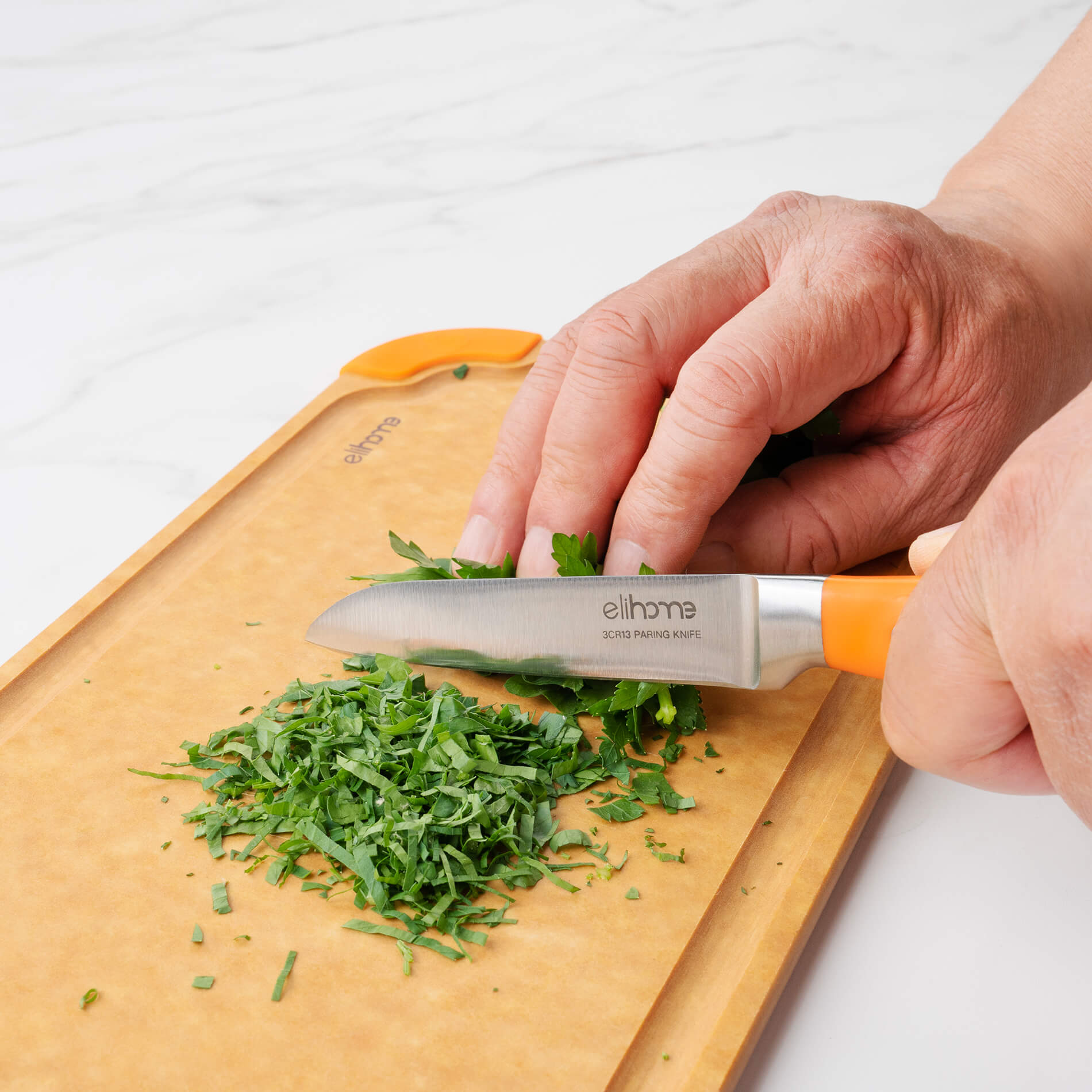 Hands using Elihome paring knife with orange handle to chop fresh herbs on wood fiber cutting board with orange silicone corners