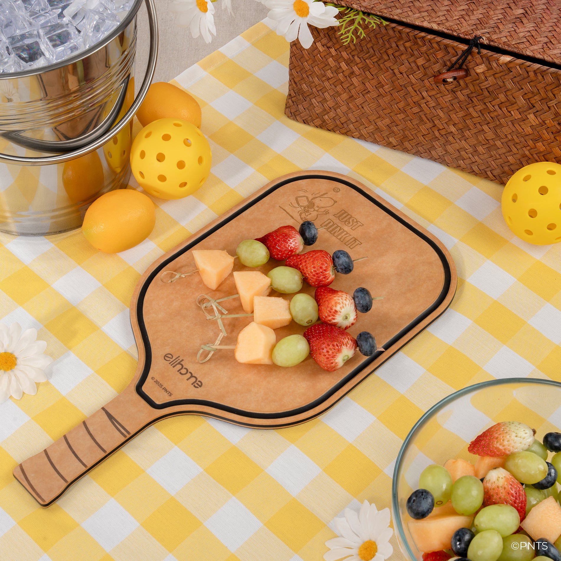 Fruit salad on a paddle-shaped cutting board with a yellow checkered tablecloth, picnic basket, and lemon on a wooden surface.