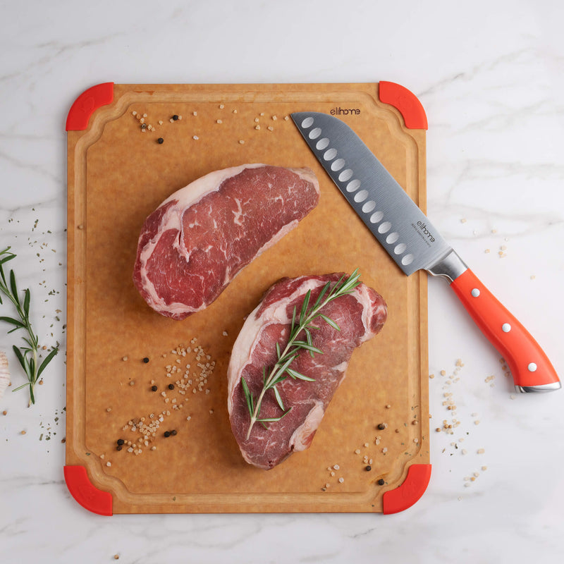 Elihome wood fiber cutting board with red silicone corners and santoku knife, displayed with raw steaks and rosemary on marble countertop