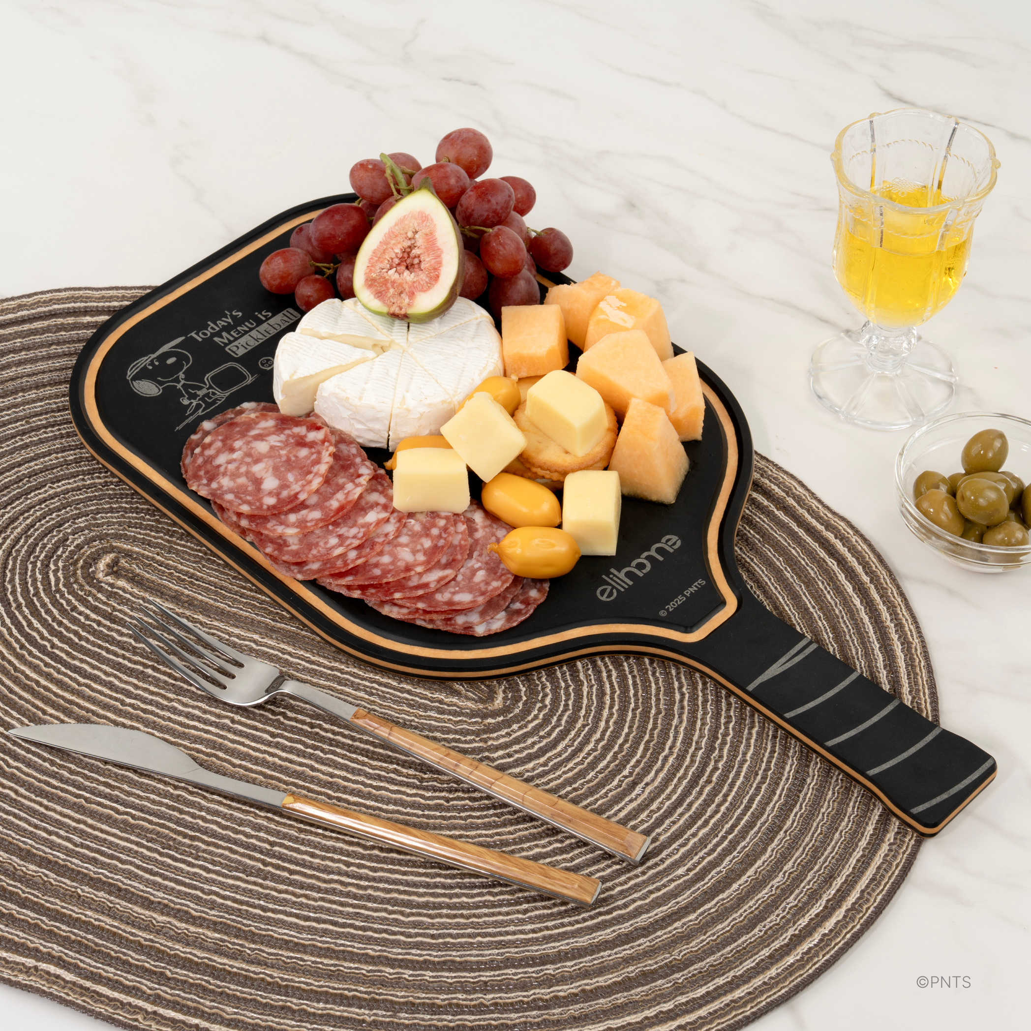 Black side of an Elihome x Peanuts pickleball paddle board engraved with Today's Menu is Pickleball, served with cheese, salami, figs and grapes