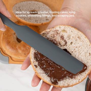 Hand using the Elihome x Peanuts Silicone Spreader to spread chocolate on a bagel half, ideal for spreading butter, frosting cakes, icing cookies, smoothing fillings and scraping jars