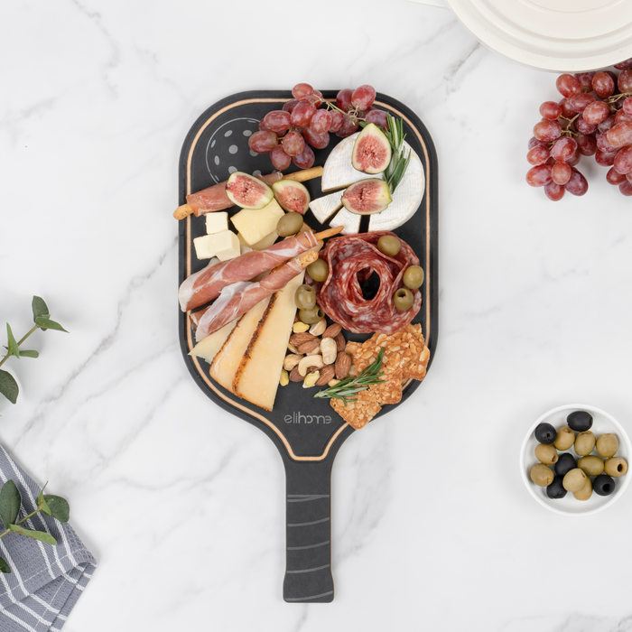 Cured meats, cheese, fresh figs, grapes, and olives arranged on the black side of an Elihome pickleball paddle serving board