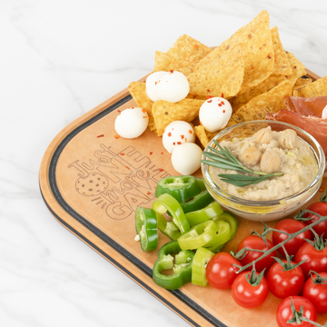 Tortilla chips, hummus, mozzarella balls, sliced peppers, and cherry tomatoes served on an Elihome pickleball paddle board engraved with Just One More Game