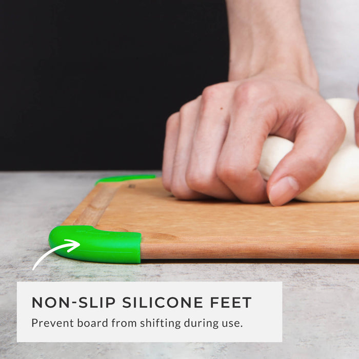 A close-up side view of a hand pressing down on the cutting board to demonstrate the stability provided by the green non-slip silicone feet gripping the countertop surface, preventing shifting during use.